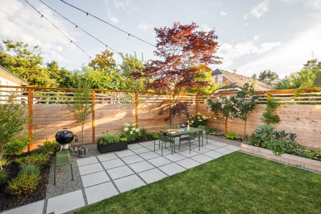 Photo of finished corner of yard with concrete paver patio, dining table and chairs, Japanese maple tree, and planting beds and containers.