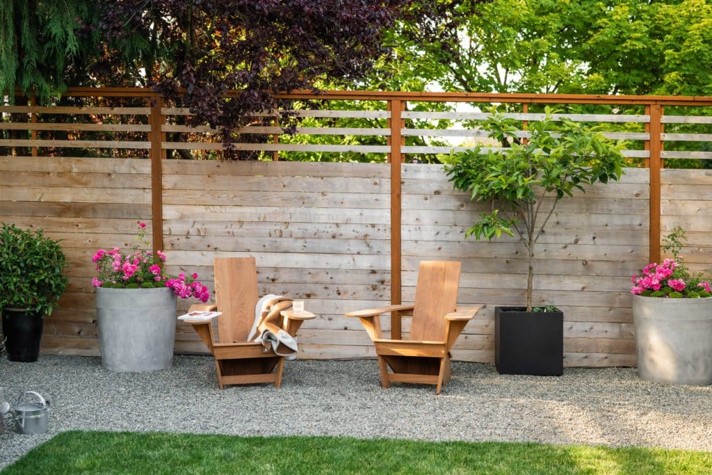 Photo of finished yard with closeup of gravel seating area with modern adirondack chairs and large planters with colorful flowers and trees.